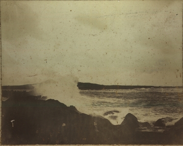 Black and white photograph with small amount of blue tint in water of a stormy sea  with large wave and rocks