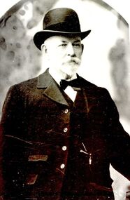 Black and white photograph from the waist up of a male with a bow tie, top hat and formal coat