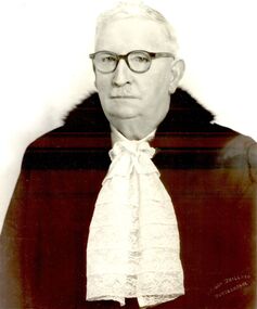 Photograph of an older male wearing glasses a black jacket with collar and a lace jabot necktie