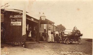 Wooden weatherboard building with, 2 children under the verandah 4 women and two men standing near car and two men in early car