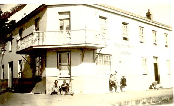 Corner of 2 story building with balcony, 2 males sitting on step and children leaning on wall