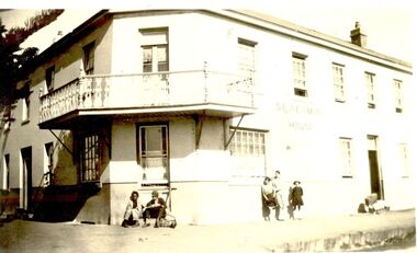 Corner of 2 story building with balcony, 2 males sitting on step and children leaning on wall