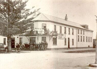 2 story rendered building with small corner balcony large group of males and females posed in front, pine tree to the left with rear of car