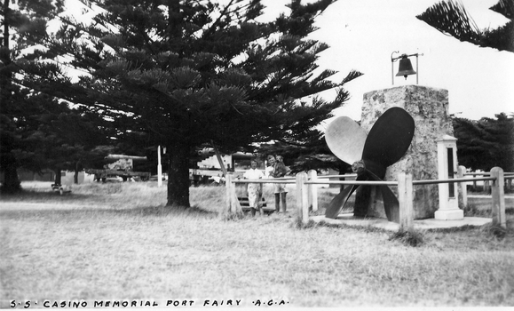 Memorial featuring ship propeller and wreck bell surrounded by a pipe fence with a couple and their dog