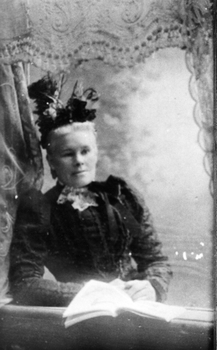 Formal portrait of woman seated with an open book and wearing a  feathered hat and lace jabot