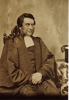 Sepia photograph of gentleman with large sideburns dressed in academic gown with white jabot 