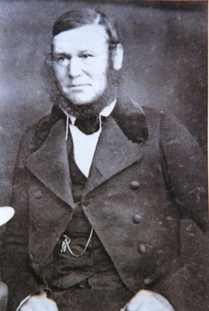 Man with hair parted on left and a full outline beard wearing a wide collared jacket and a watch chain is visible on his chest