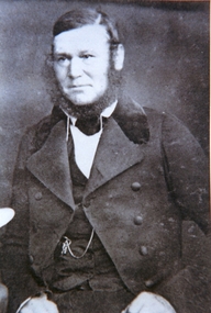 Man with hair parted on left and a full outline beard wearing a wide collared jacket and a watch chain is visible on his chest