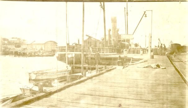 S.s.Casino berthed at wharf small dinghies in foreground two males in middle ground, lifeboat half round shed to the left and  train carriages in distance on right 