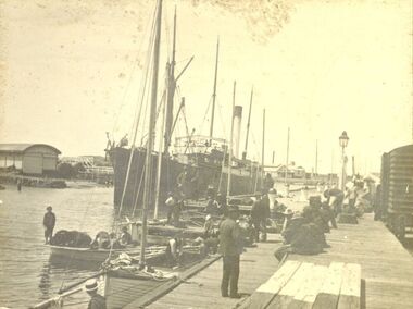 Ship at Belfast and Koroit Steam Navigation wharf with couta boats in foreground and fishermen on board and on the wharf, lifeboat half round shed on the left, train carriage on left and in background on the left the Rabbit factory