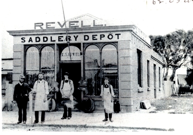 Black and white photograph of a detached shop with two high arched windows either side of the door goods on footpath and 4 males with aprons on on footpath