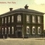 Coloured photograph of the double storied bluestone “ivy” Bank of Australasia with hitching posts either side of corner front door an electricity pole to the left a male, three children and two dogs to the right
