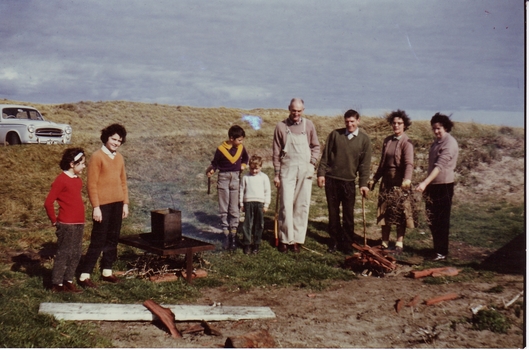 Group of people with a kerosine tin on a fire with another to the right being set dunes in the background white car on left
