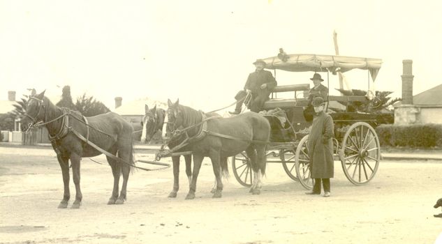 Horse drawn dray with seats for passengers around sides and drop down canvas blinds.3 horses harnessed and 3 males observing