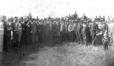 Large semi circle of males formally dressed with male in the centre holding a spade to turn the first sod.