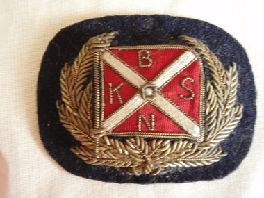 rectangular hat badge with gold laurel leaves embracing the red and white flag of the Belfast and Koroit Steam Navigation Company