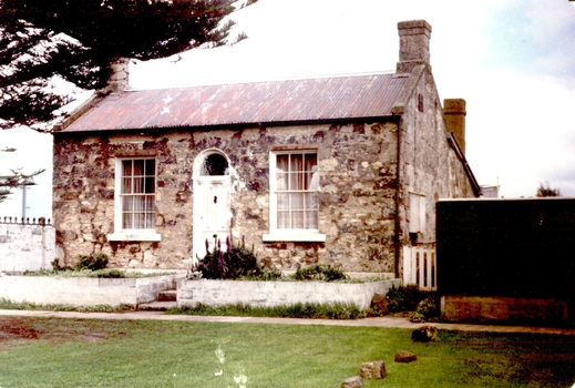 Bluestone and limestone cottage with arched doorway and windows either side. A stone fence around the front