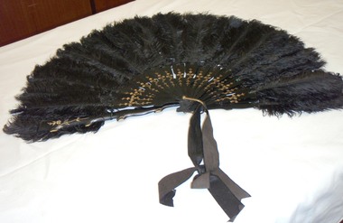 A large fan made from early plastic and feathers with a repeated gold floral design on each slat held together with a brass loop
