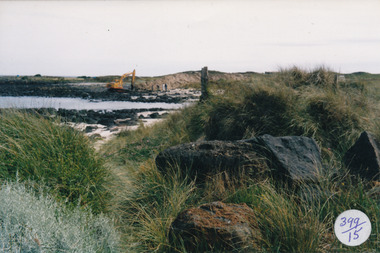 Looking through marram grass across rocks sand and water at a bright yellow digger in centre background 