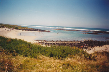 Coloured photograph of pea soup beach with the reef in the background with people in the water and on the sand
