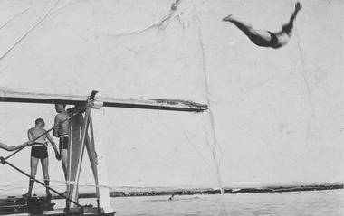 Black and white photograph of the diving board with 2 males standing  and one diving