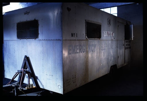 Silver metal trailer with two rectangular windows; body of the trailer is riveted together and painted with the words "PMG/EMERGENCY TELEPHONE EXCHANGE"