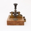 Metal device mounted on block of wood with handle that moves up and down via a spring.