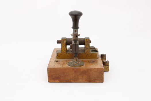 Metal device mounted on block of wood with handle that moves up and down via a spring.