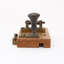 Metal device mounted on block of wood with handle that moves up and down via a spring.