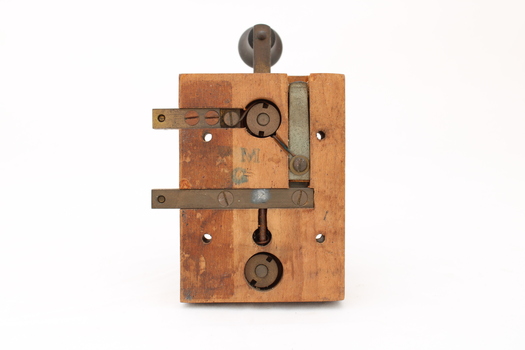 Metal device mounted on block of wood with handle that moves up and down via a spring.