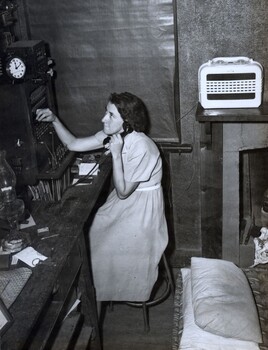 Young woman sitting on chair holding a telephone receiver to her ear while plugging a cord into a switchboard; pillows in the foreground.