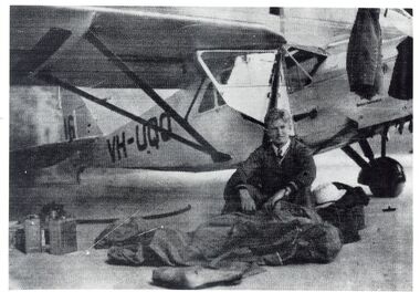 Young man in a shirt, tie and possible flight suit sitting in front of a high-wing monoplane with textile bags and containers in the foreground.