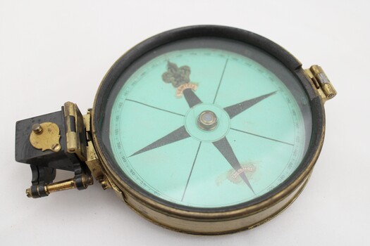 Gold coloured metal compass case with engraved lid; compass face is green printed paper with glass top.