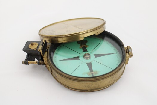 Gold coloured metal compass case with engraved lid; compass face is green printed paper with glass top.