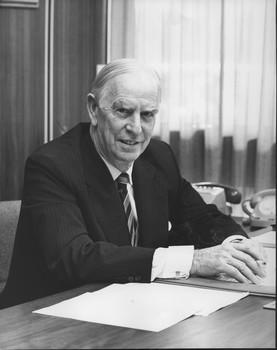 Man seated at desk with a pen in his hand, telephones to the side and a piece of paper on the desk