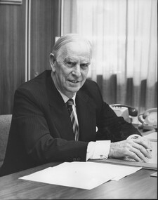 Man seated at desk with a pen in his hand, telephones to the side and a piece of paper on the desk