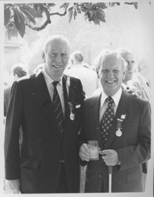 Two suited men smile as they face the camera, one holds a glass and a white cane