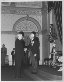 Suited man shakes hands with man dressed in dressed in tails and wearing a row of medals