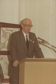Man in suit with glasses looks towards the audience from a podium