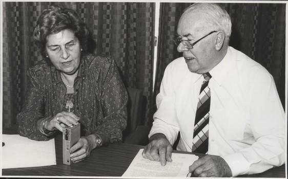 Woman holding tape recorder speaks with older man who has typed pages before him