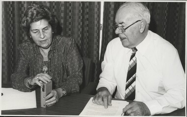 Woman holding tape recorder speaks with older man who has typed pages before him