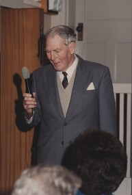 Older man standing with microphone in hand, before an audience