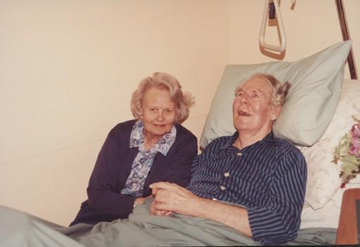 John Blanch reclining in nursing home bed with wife (?) beside him