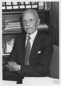 Man in suit and tie crosses his arms and smiles towards camera from behind a desk