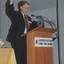 Robin Pleydell holds up a copy of the Annual Report whilst standing at the podium on stage