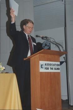 Robin Pleydell holds up a copy of the Annual Report whilst standing at the podium on stage