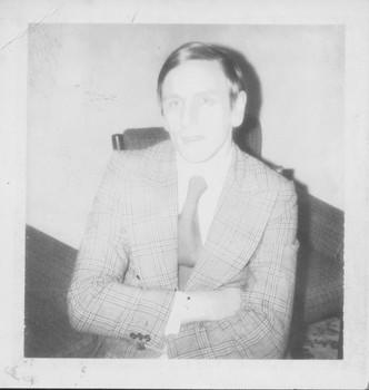 Man in plaid suit crosses his arms as he sits