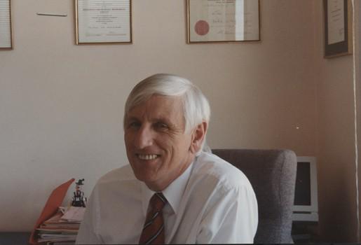 John Cook sitting in his office chair with framed certificates behind him