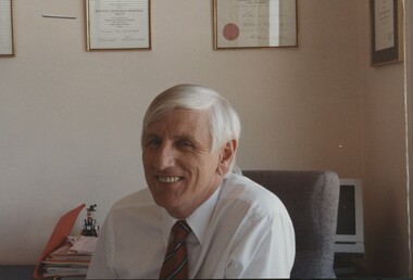 John Cook sitting in his office chair with framed certificates behind him