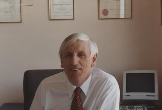 John Cook sitting in his office chair with framed certificates behind him
